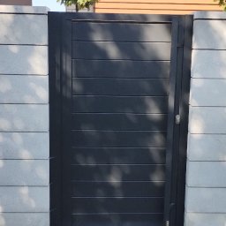 MD Nowoczesne konstrukcje - Nowoczesna, grafitowa furtka z poziomymi panelami, wkomponowana w szary, betonowy mur. W tle widoczny fragment domu z drewnianą elewacją i zielone drzewko.