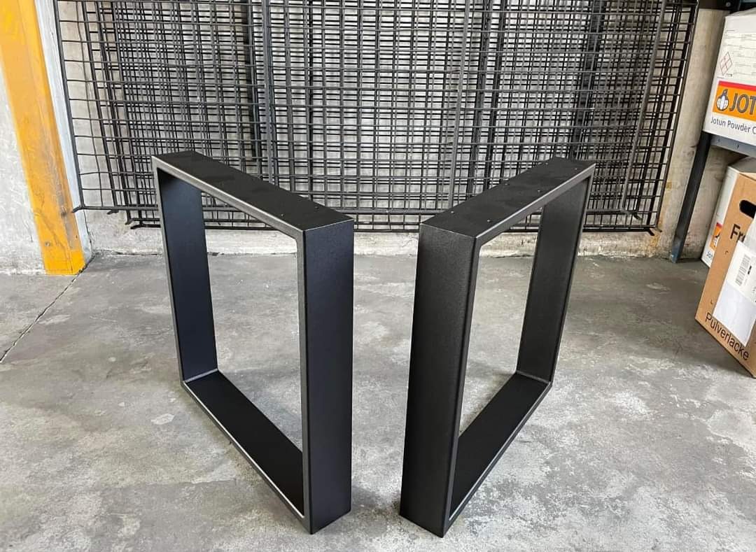 Dwie czarne, metalowe nogi do stołu w kształcie prostokąta, ustawione na szarym betonie, z metalową siatką i kartonami w tle. Widoczne ślady spawania i łączenia.