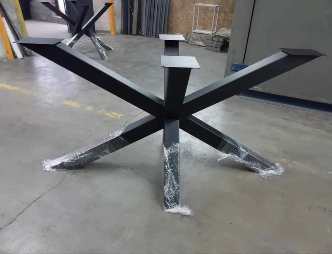 Surowa, czarna, metalowa podstawa stołu o geometrycznym kształcie X, z elementami zabezpieczonymi folią, na szarym, betonowym podłożu w warsztacie.