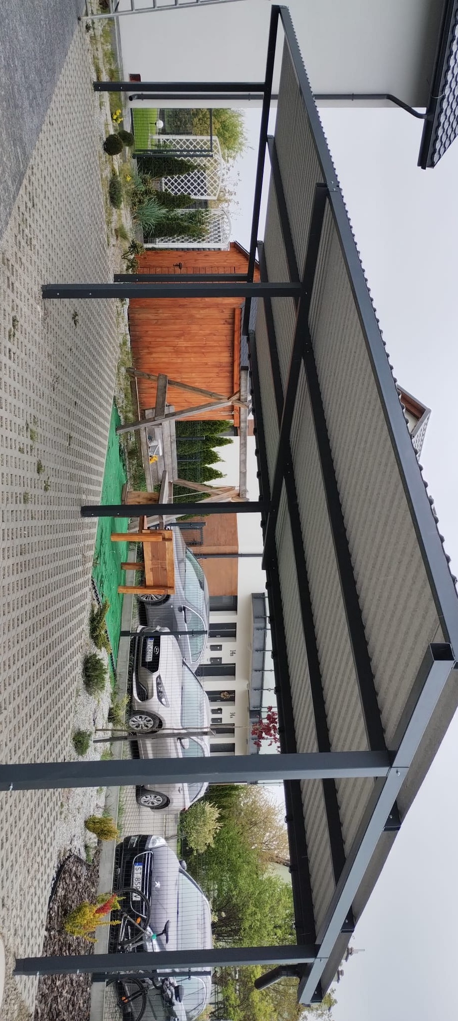 Nowoczesna, szara pergola metalowa z zadaszeniem, wsparta na solidnych słupach, otoczona kostką brukową i zielenią. W tle widoczne zaparkowane samochody i zabudowa mieszkalna.