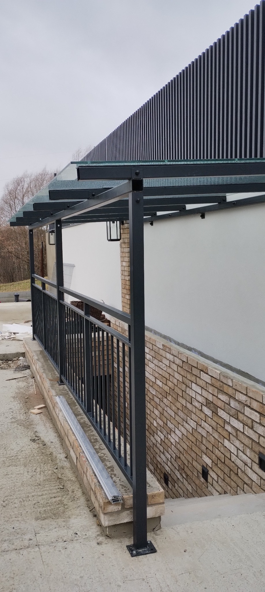 Nowoczesna, metalowa pergola z balustradą i szklanym dachem, z widocznymi elementami montażowymi, na tle ceglanej ściany i białej elewacji z pionowymi panelami.