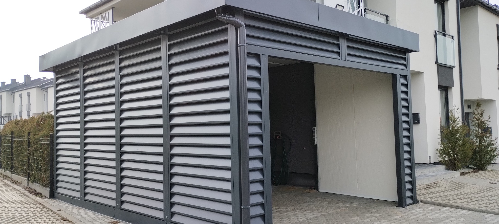 Nowoczesna, szara pergola z lamelami, przylegająca do budynku. Widoczny fragment posesji i elewacja domu. Charakterystyczne poziome lamele.