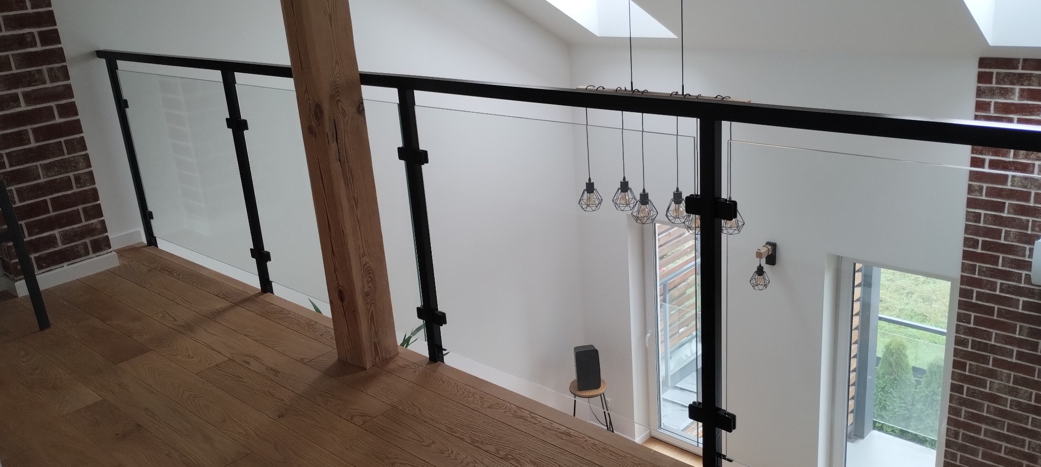 Nowoczesna balustrada szklana z czarnymi metalowymi słupkami i drewnianym filarem, widok z góry na drewnianą podłogę i jasne wnętrze.