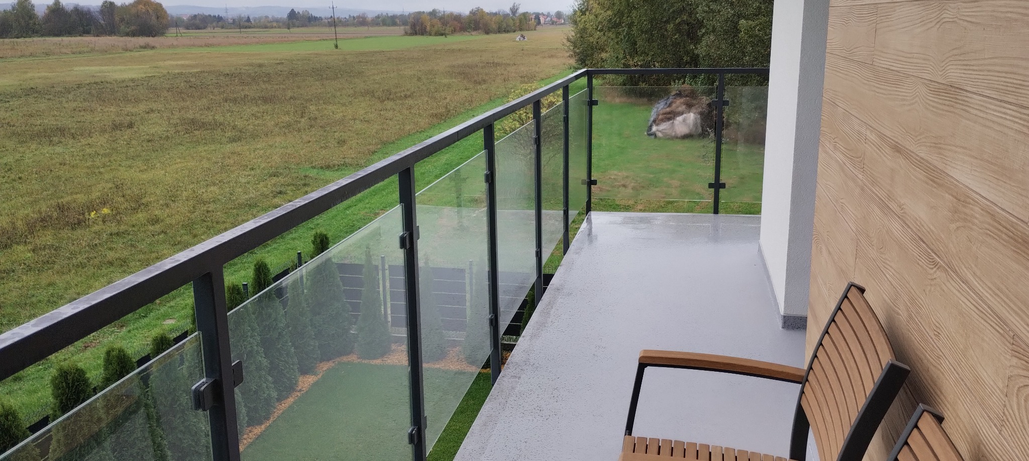 Nowoczesny balkon z szarą podłogą, drewnianą ścianą i szklaną balustradą z ciemną ramą. W tle widok na zielone pole i drzewa. Część krzesła z drewnianym oparciem.