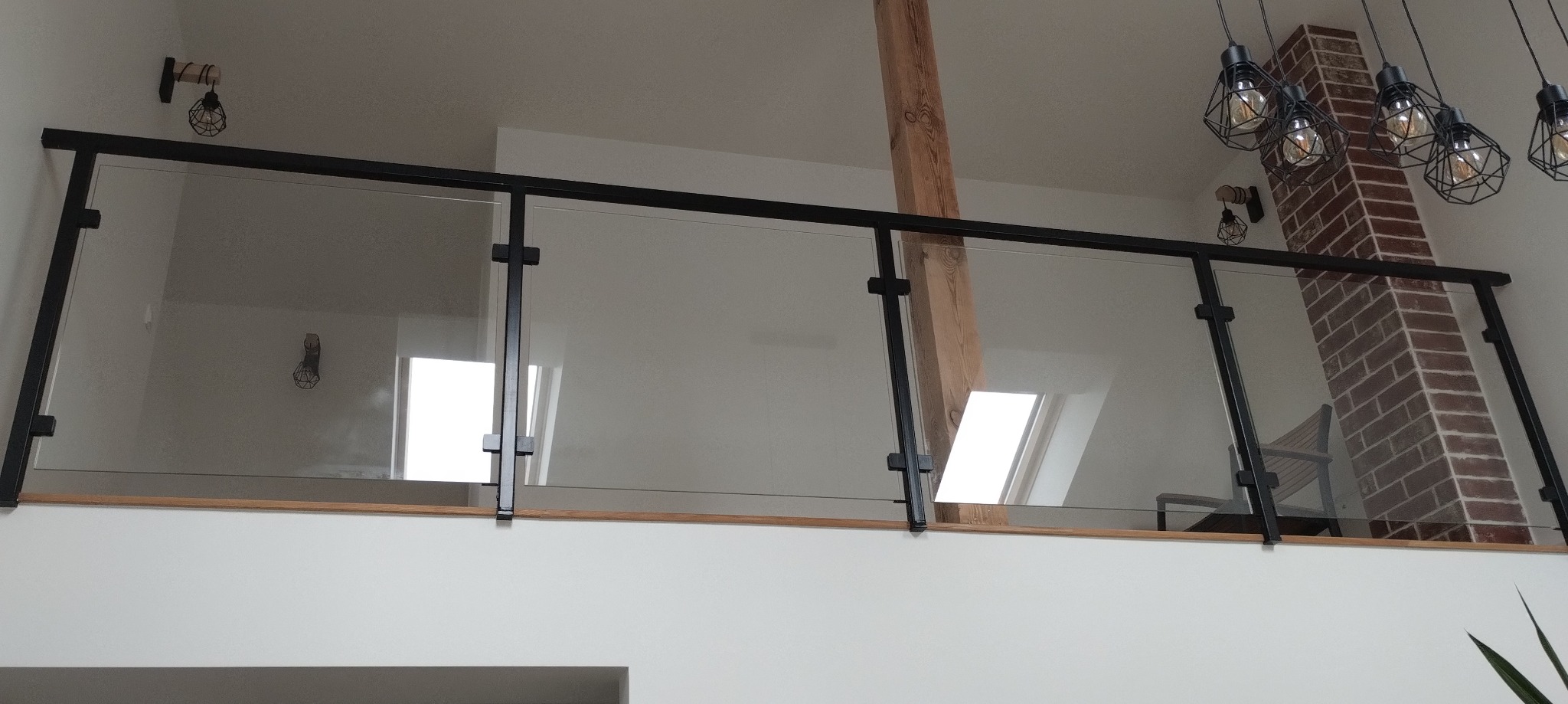 Szklana balustrada z czarnymi profilami na antresoli, widok z dołu. W tle ceglany komin i designerskie lampy, oświetlające wnętrze domu.