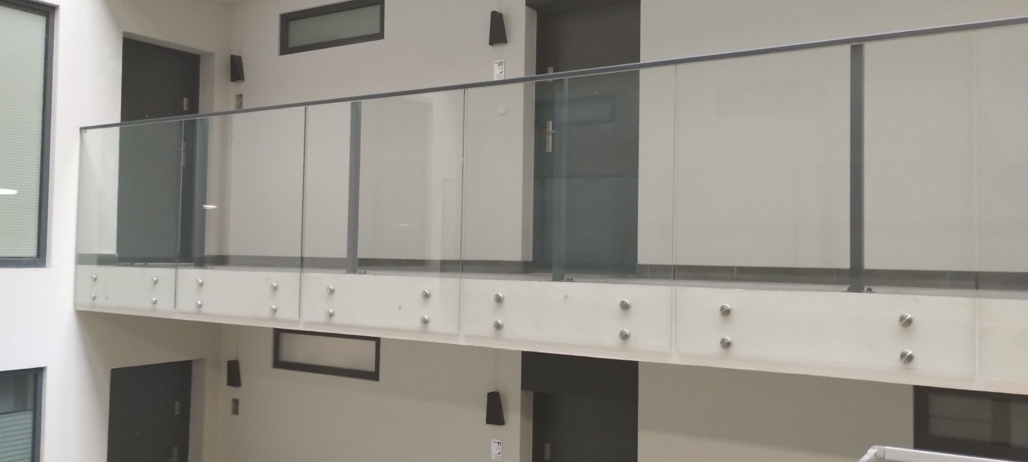 Szklana balustrada z metalowymi elementami mocującymi, widoczna z perspektywy dolnej kondygnacji, w nowoczesnym wnętrzu budynku z drzwiami i oknami.