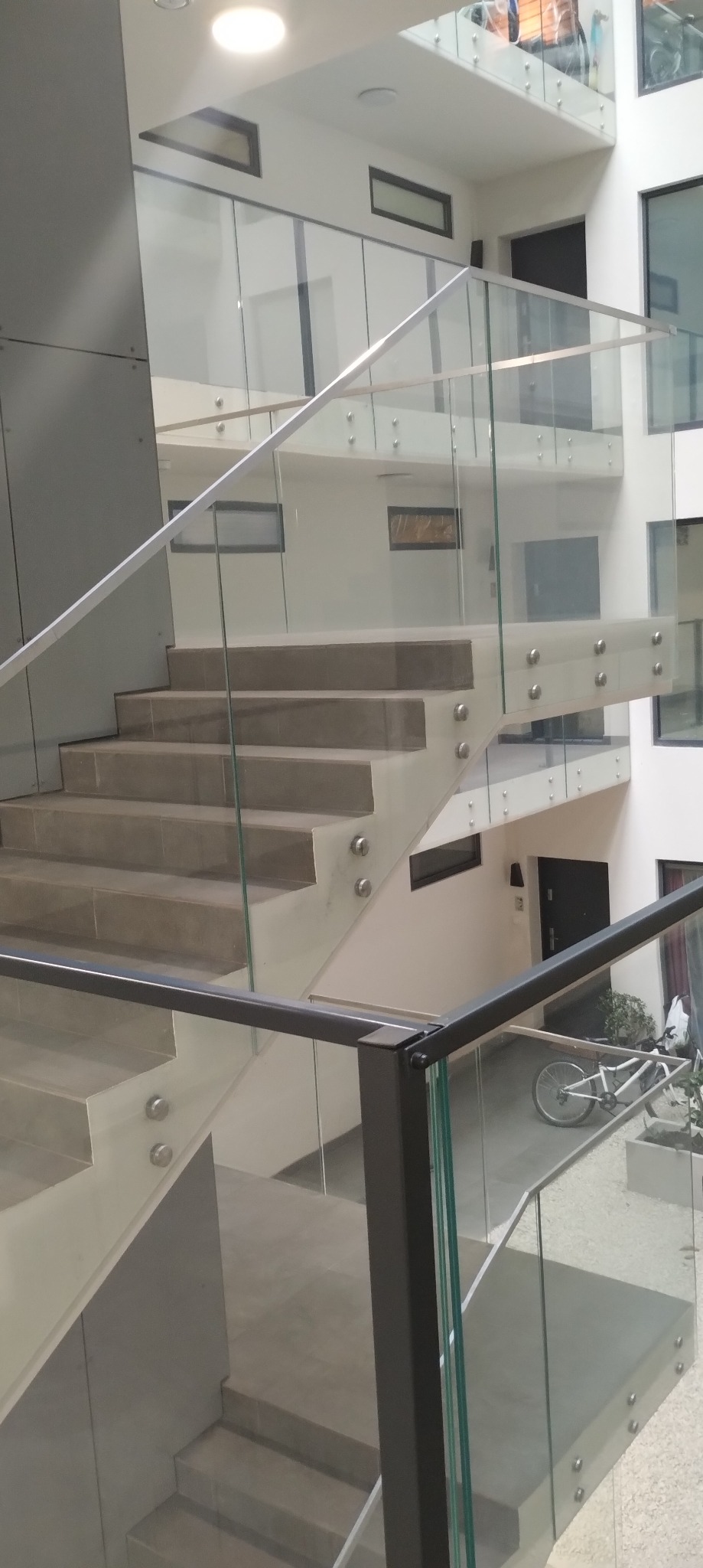 Nowoczesne schody z jasnymi stopniami i szklaną balustradą, mocowaną punktowo. W tle widoczny rower i jasne wnętrze budynku.