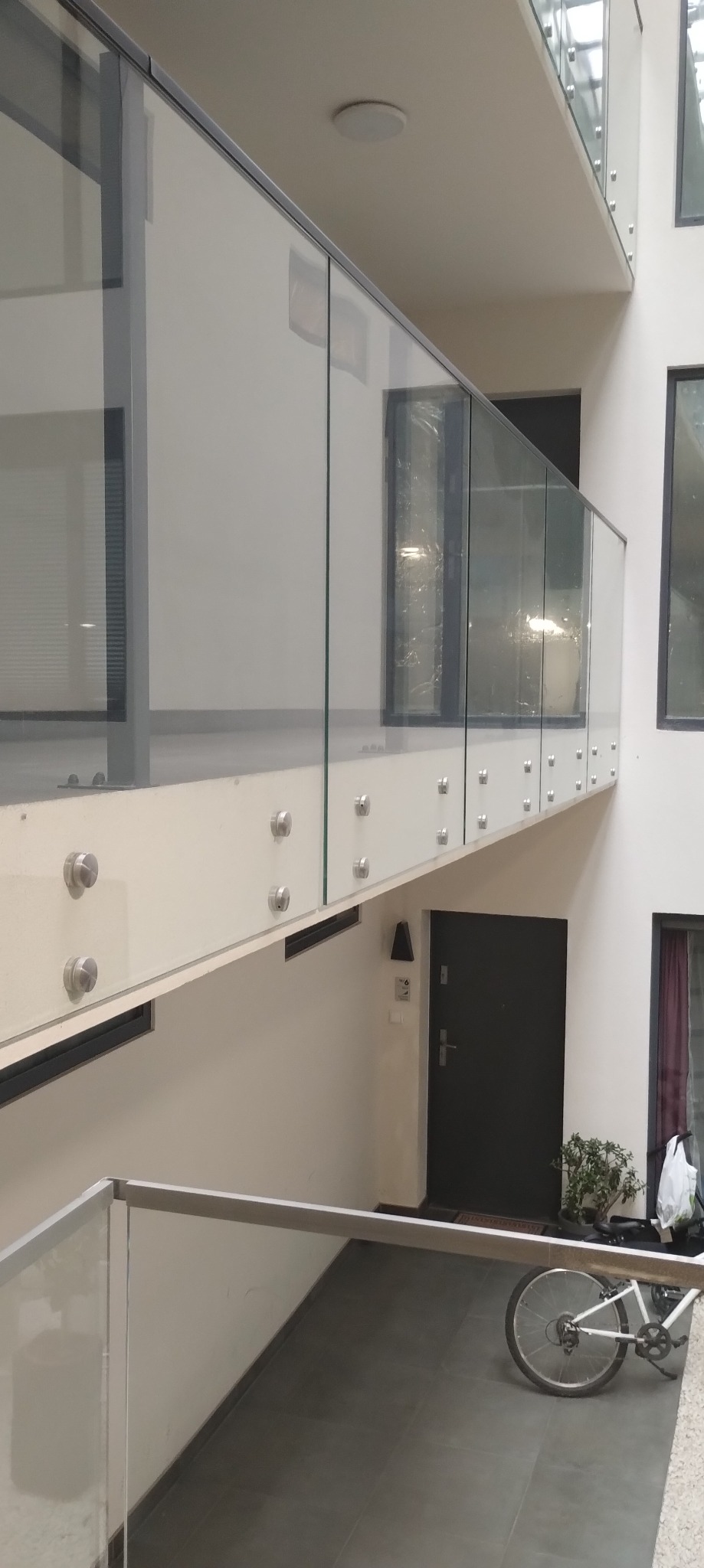 Szklana balustrada z mocowaniami punktowymi w nowoczesnym wnętrzu. Widoczny rower i drzwi wejściowe w tle. Jasne, neutralne kolory ścian i podłogi.