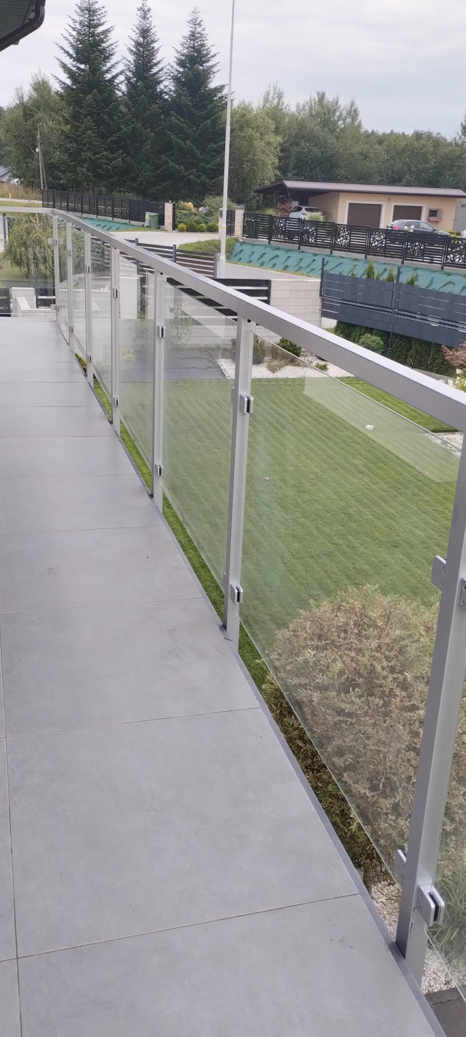 Nowoczesna balustrada szklana z aluminiowymi słupkami na balkonie z szarymi płytkami. Widok na zadbany trawnik i dom z ciemnym ogrodzeniem.