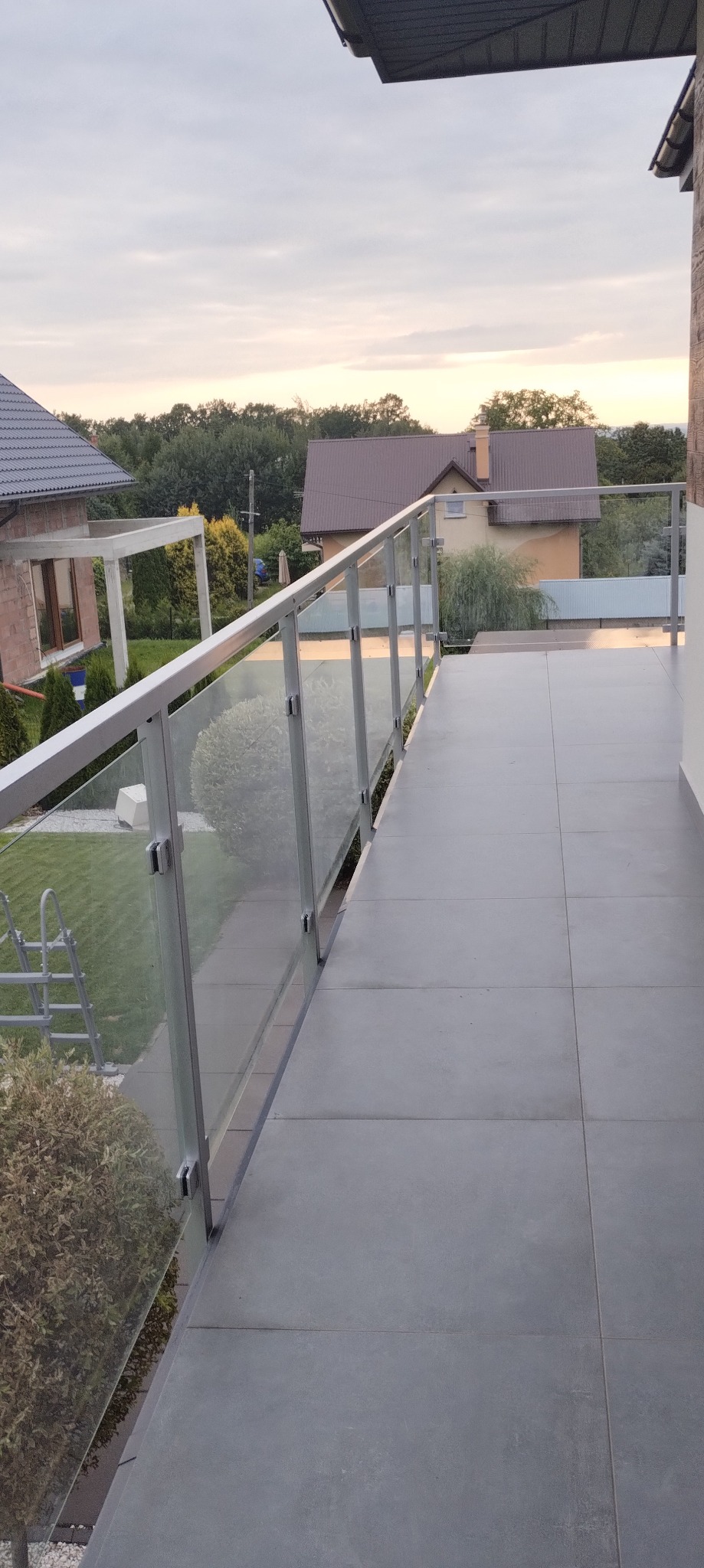 Nowoczesny balkon z szarą, kwadratową podłogą i szklaną balustradą z aluminiowymi profilami. W tle domy i zieleń. Widok z góry, spokojne oświetlenie.