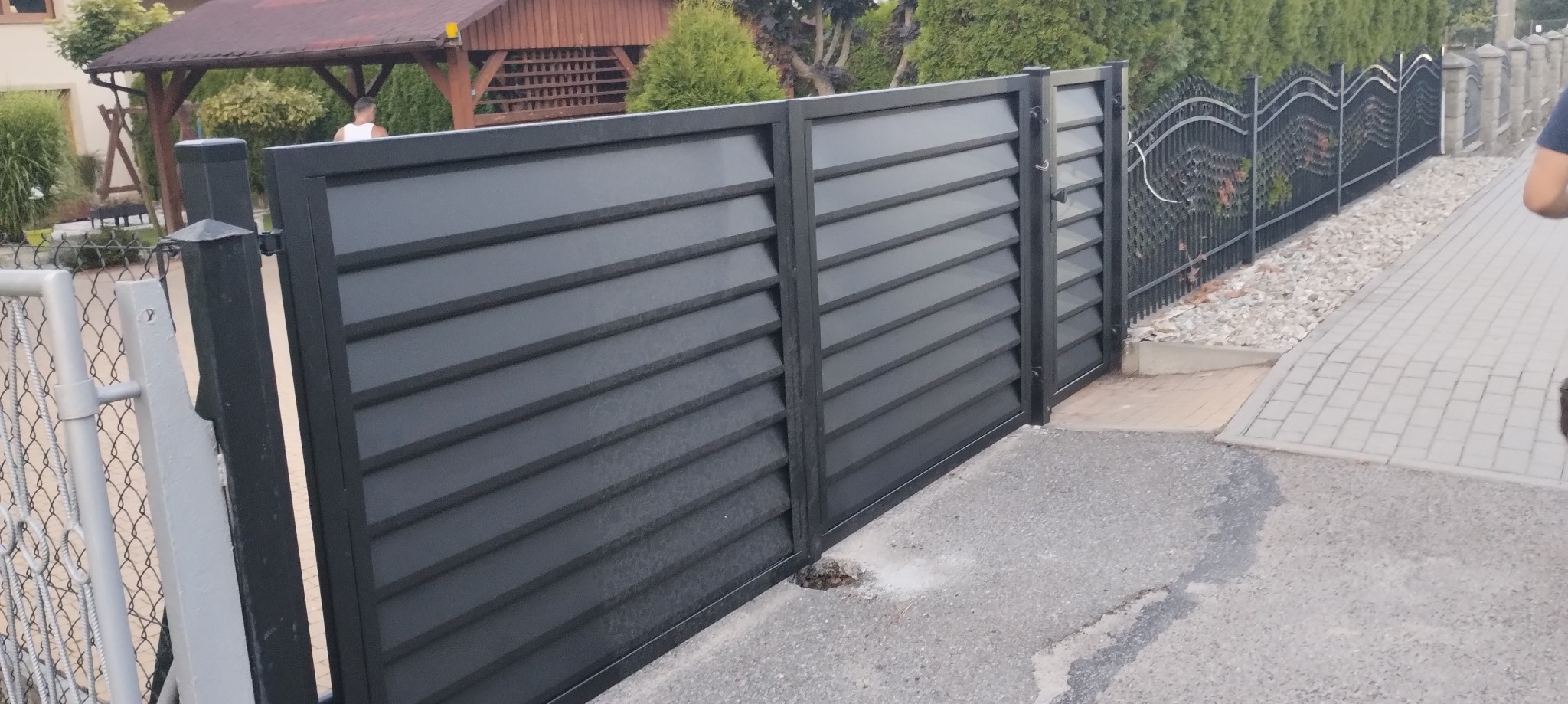 Nowoczesna, grafitowa brama wjazdowa z poziomymi lamelami, uzupełniona o klasyczne, metalowe ogrodzenie z ozdobnymi elementami. Widoczne detale montażowe.