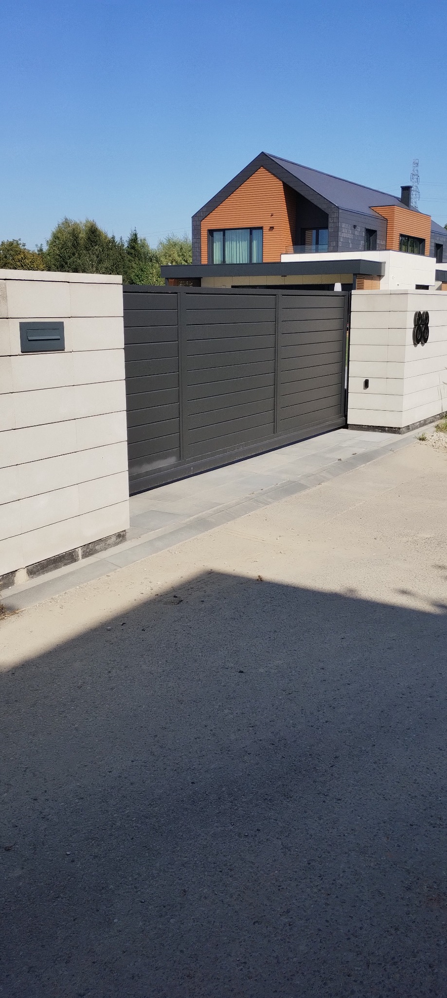 Nowoczesna, przesuwna brama wjazdowa z poziomych paneli w kolorze grafitowym, osadzona między dwoma jasnymi, murowanymi filarami. W tle widoczny dom z elementami drewnianymi.