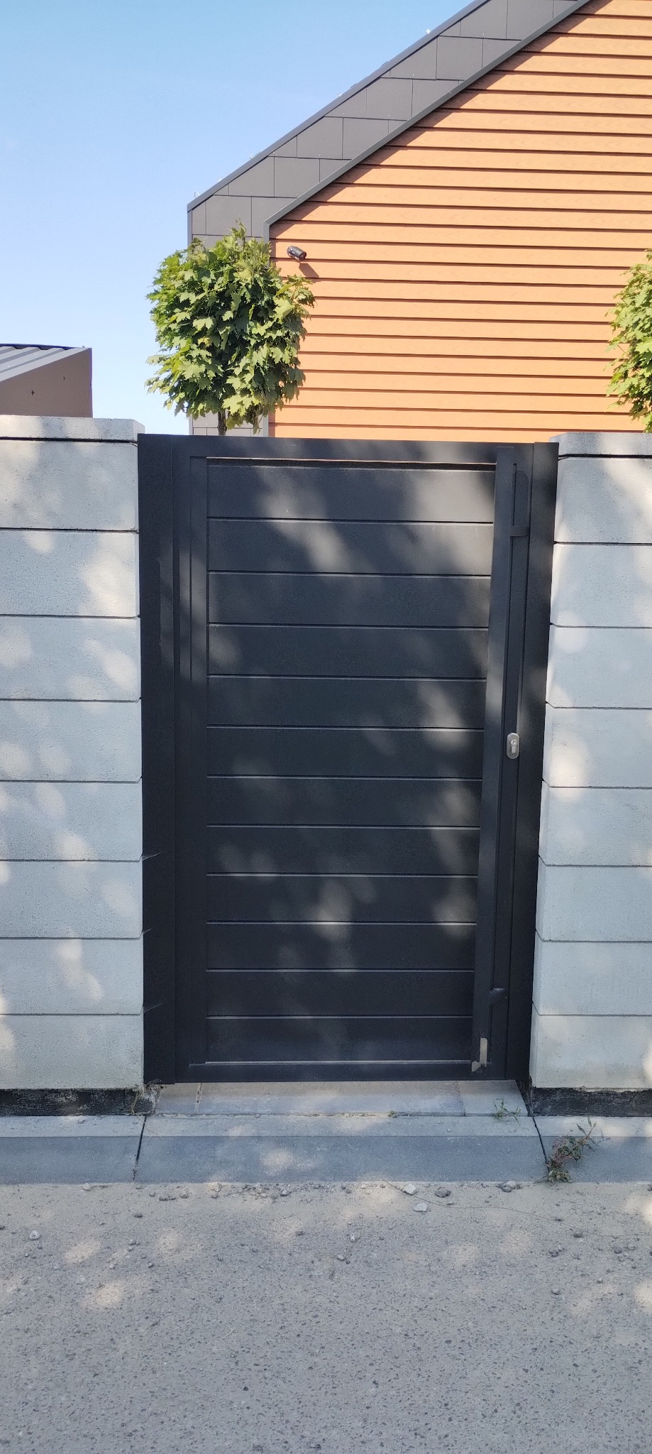 Nowoczesna, grafitowa furtka z poziomymi panelami, wkomponowana w szary, betonowy mur. W tle widoczny fragment domu z drewnianą elewacją i zielone drzewko.