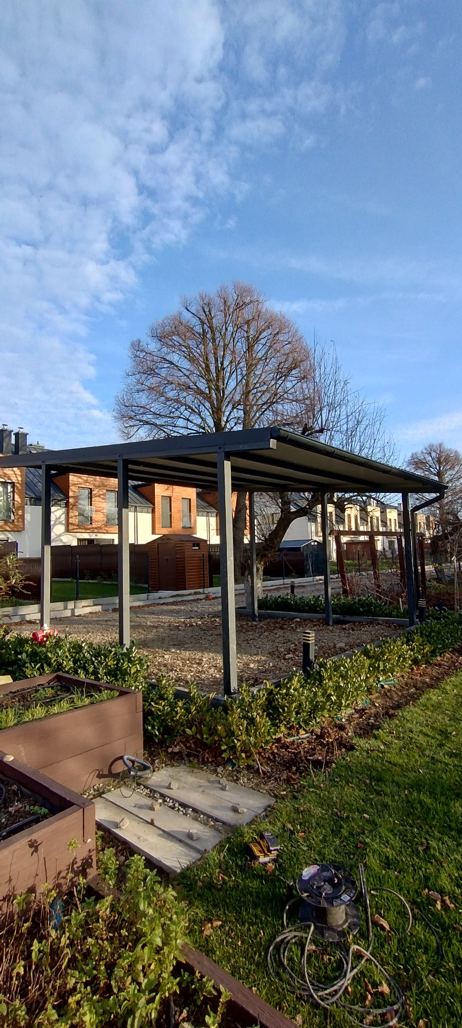 Nowoczesna, grafitowa pergola metalowa w ogrodzie, z widocznymi narzędziami na trawie. W tle domy jednorodzinne i błękitne niebo z chmurami.