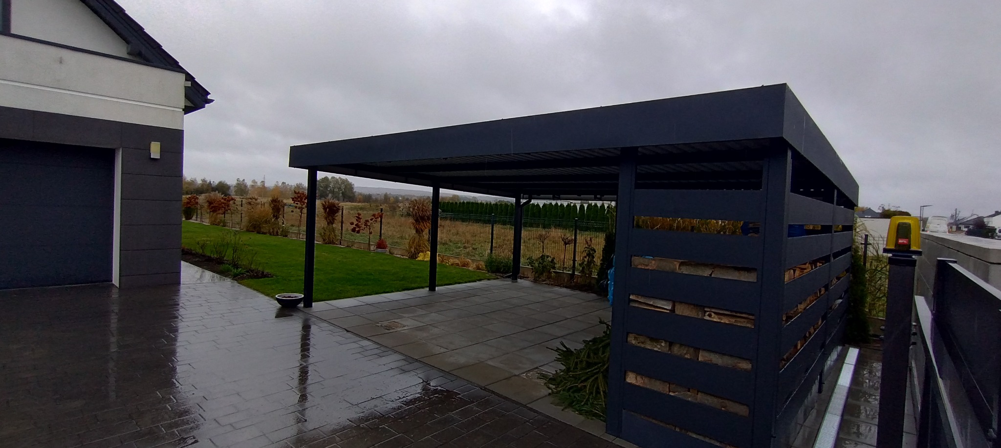 Nowoczesna, grafitowa pergola metalowa z ażurową ścianą boczną, na tle posesji z zielonym trawnikiem i kostką brukową. Widok na jesienny krajobraz w tle.