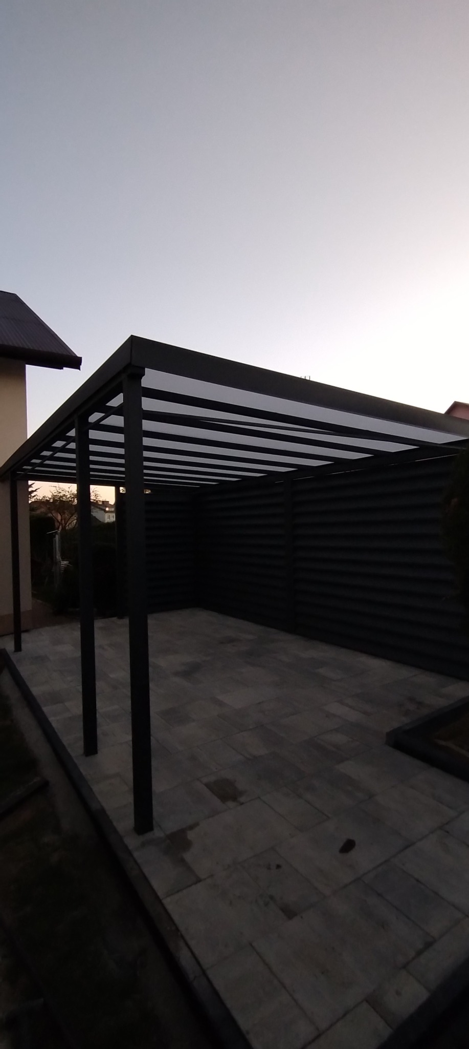 Nowoczesna, czarna pergola metalowa z zadaszeniem lamelowym, na tle szarej ściany i kostki brukowej. Minimalistyczny design, wieczorny kadr.