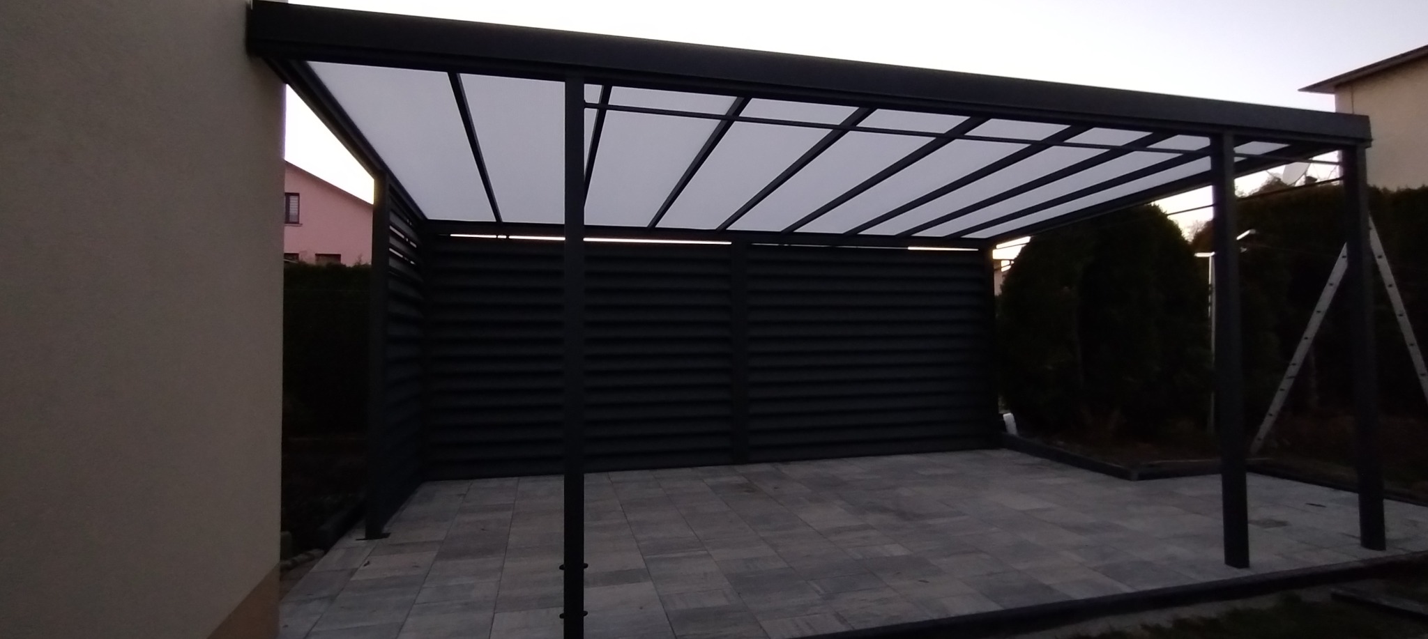 Nowoczesna, grafitowa pergola z zadaszeniem z poliwęglanu, przylegająca do budynku. Widoczne poziome lamele w tylnej ścianie i szara kostka brukowa na podłożu.