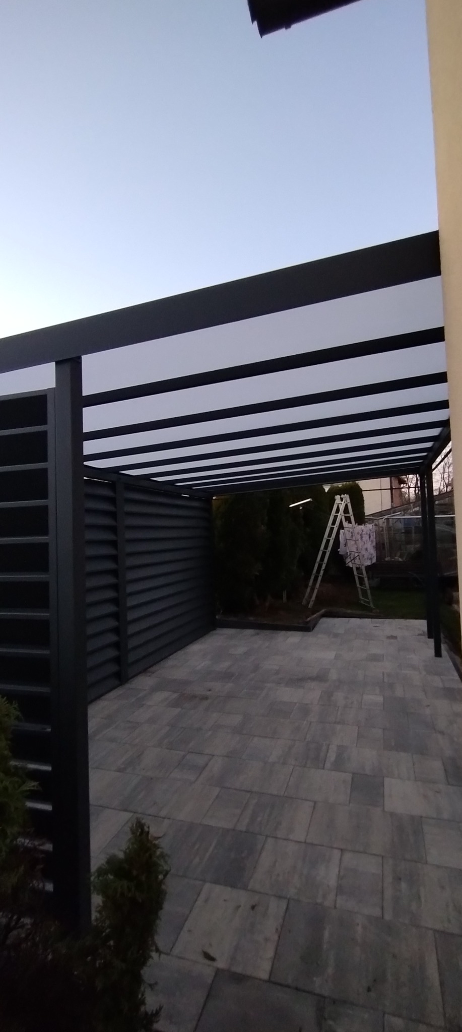 Nowoczesna pergola z ciemnymi elementami konstrukcyjnymi i jasnym zadaszeniem, widoczna z boku, na tle kostki brukowej i zieleni. W tle drabina.