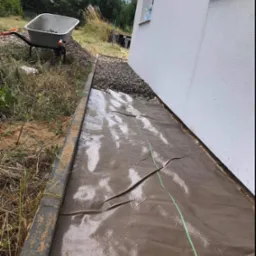 Przygotowanie podłoża pod trawnik: rozłożona siatka wzmacniająca z zielonymi liniami, obok budynku, z krawężnikiem i taczką z kamieniami w tle.