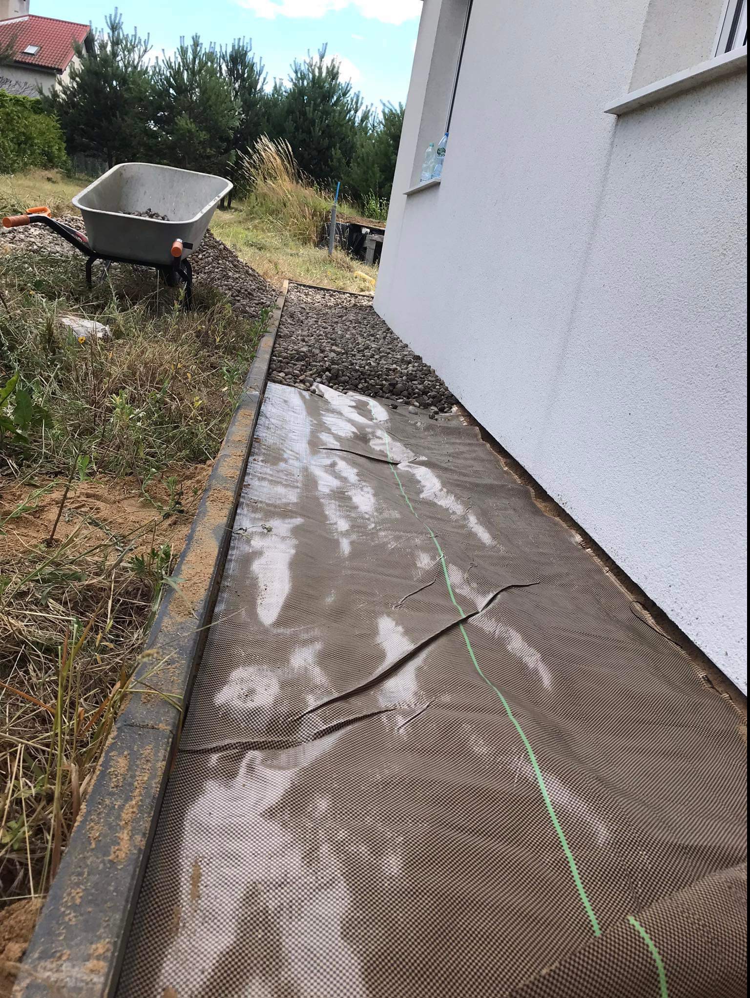 Przygotowanie podłoża pod trawnik: rozłożona siatka wzmacniająca z zielonymi liniami, obok budynku, z krawężnikiem i taczką z kamieniami w tle.