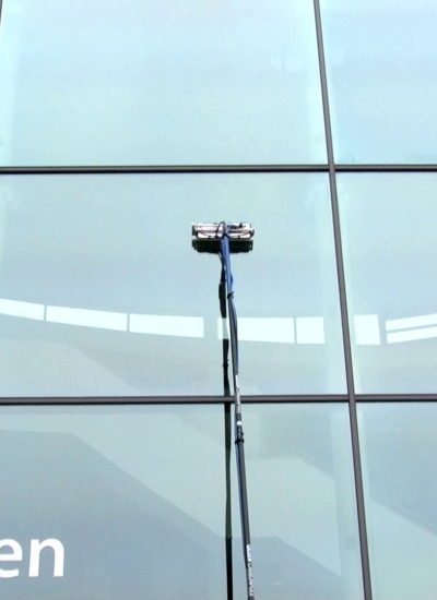 Szczotka na teleskopowym kiju podczas czyszczenia szklanej elewacji budynku, widoczne odbicia w szybach.