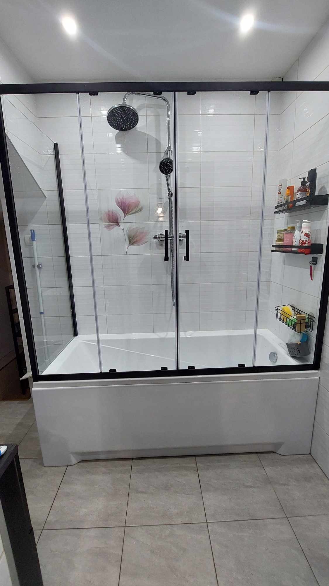 Zamontowana wanna z czarną ramą parawanu, deszczownicą i dodatkową słuchawką, białe płytki z motywem kwiatowym oraz półka z kosmetykami.