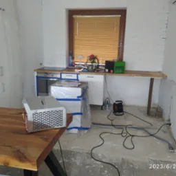 Osuszanie po zalaniu: widoczny osuszacz kondensacyjny na drewnianym stole w pomieszczeniu z zabezpieczonymi folią meblami kuchennymi i odkrytą posadzką.