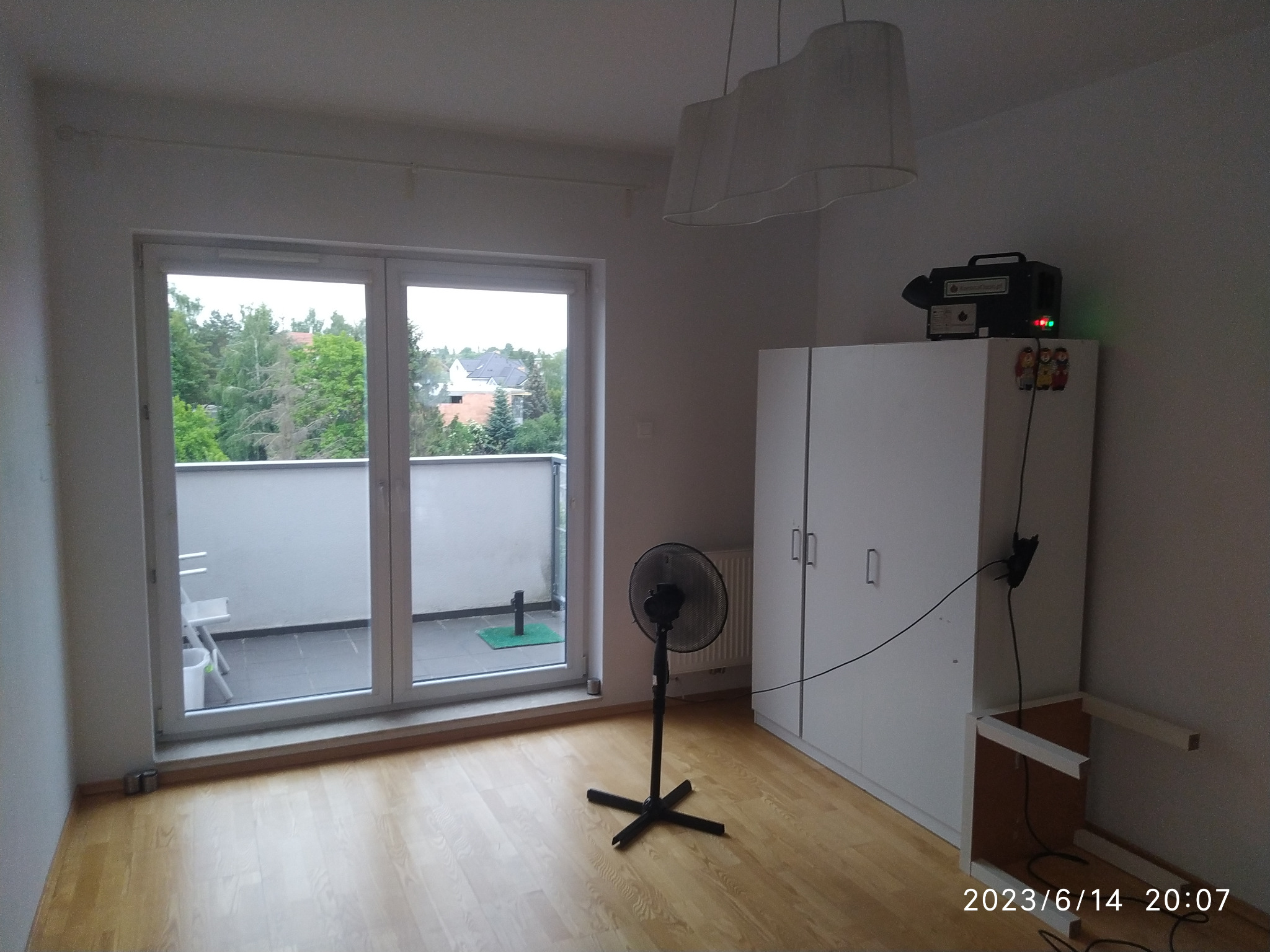 Pokój z parkietem, wentylatorem stojącym i urządzeniem do osuszania na szafie, widok na balkon i zieleń za oknem.