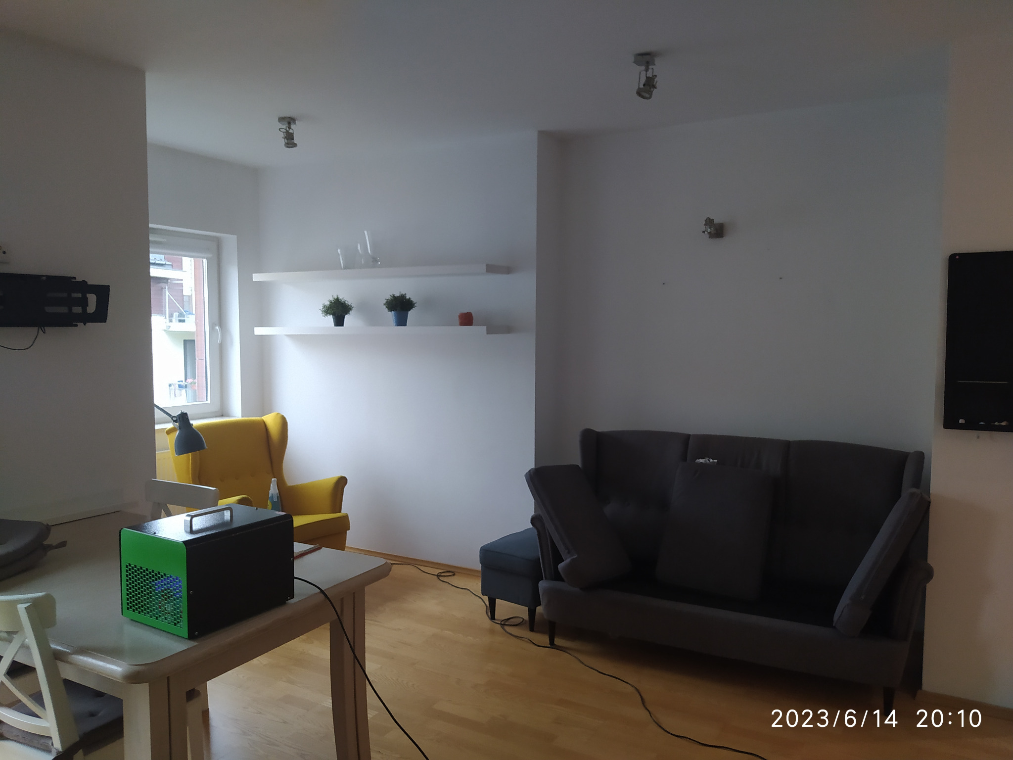 Czarny osuszacz powietrza z zielonymi bokami stoi na stole w salonie, widoczny szary fotel i żółty fotel w tle.