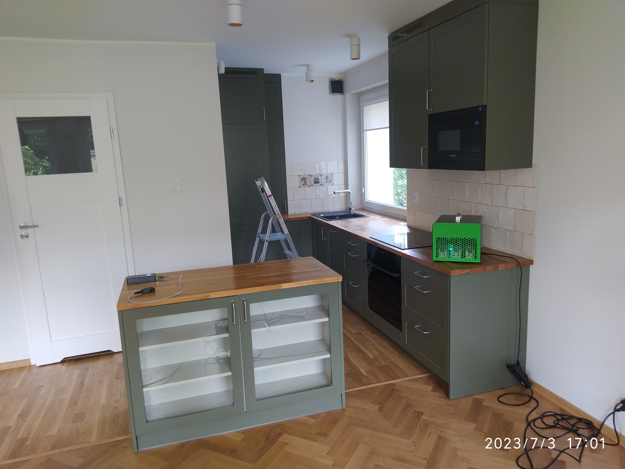 Nowoczesna kuchnia w odcieniach zieleni z drewnianym blatem, widoczny ozonator powietrza na blacie, drabina oparta o ścianę, parkiet w jodełkę.