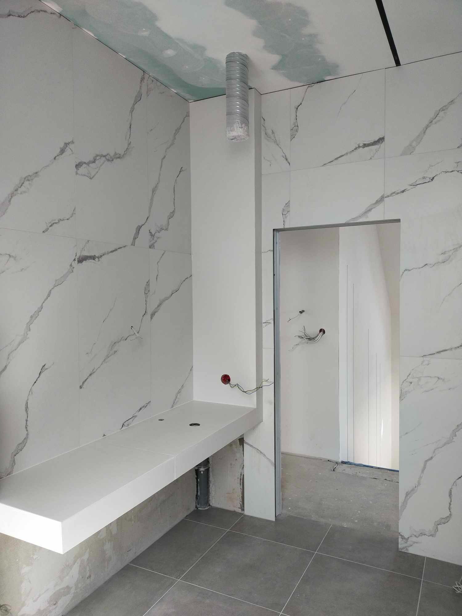 Łazienka w trakcie remontu: białe płytki imitujące marmur na ścianach, szara podłoga z płytek, biały blat pod umywalkę, widoczne przewody elektryczne i rura wentylacyjna pod sufitem.
