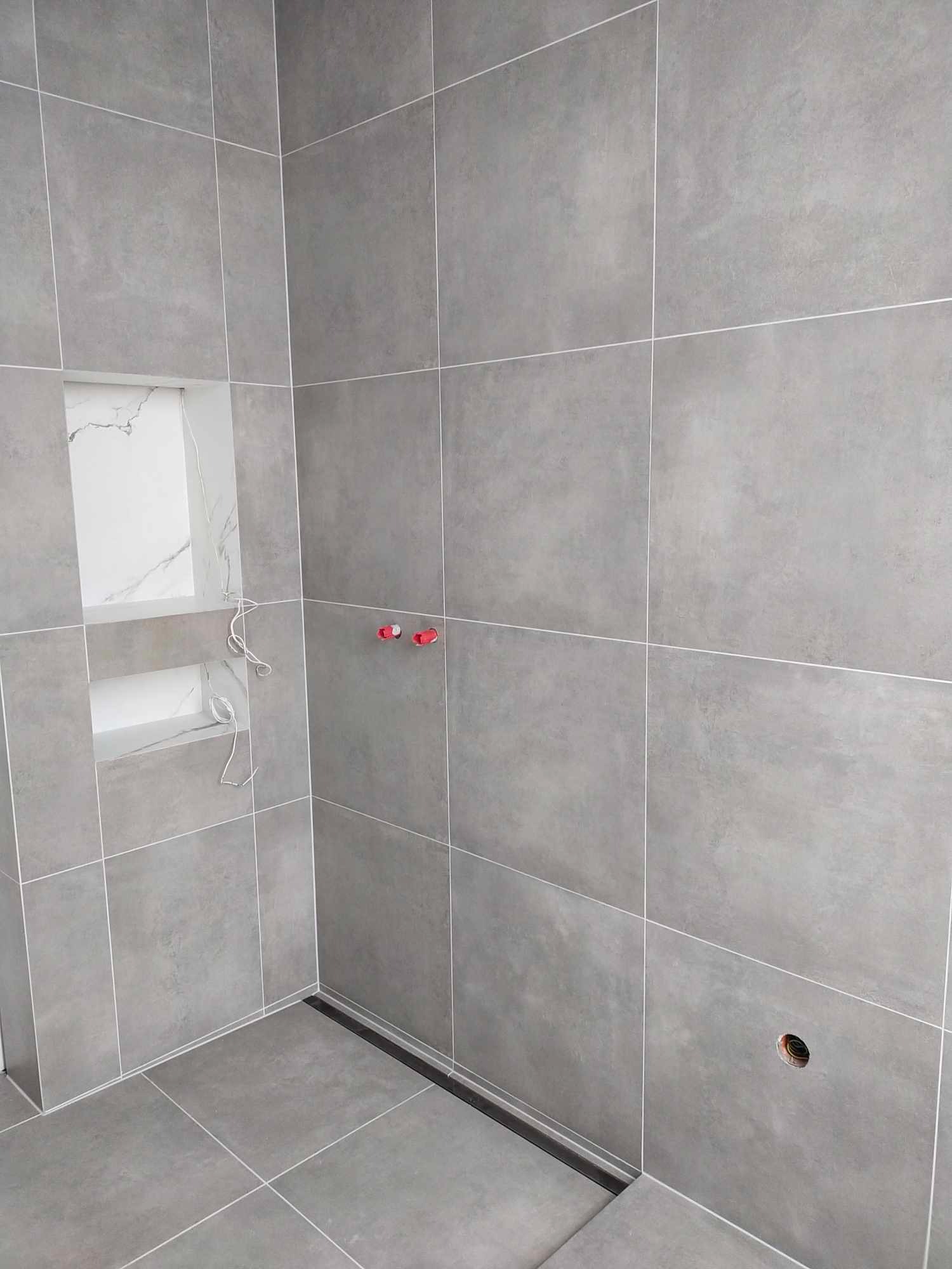 Szare, matowe płytki wielkoformatowe na ścianach i podłodze w kabinie prysznicowej. W ścianie wnęki wyłożone białymi płytkami z marmurowym wzorem. Widoczny odpływ liniowy i wystające rurki...
