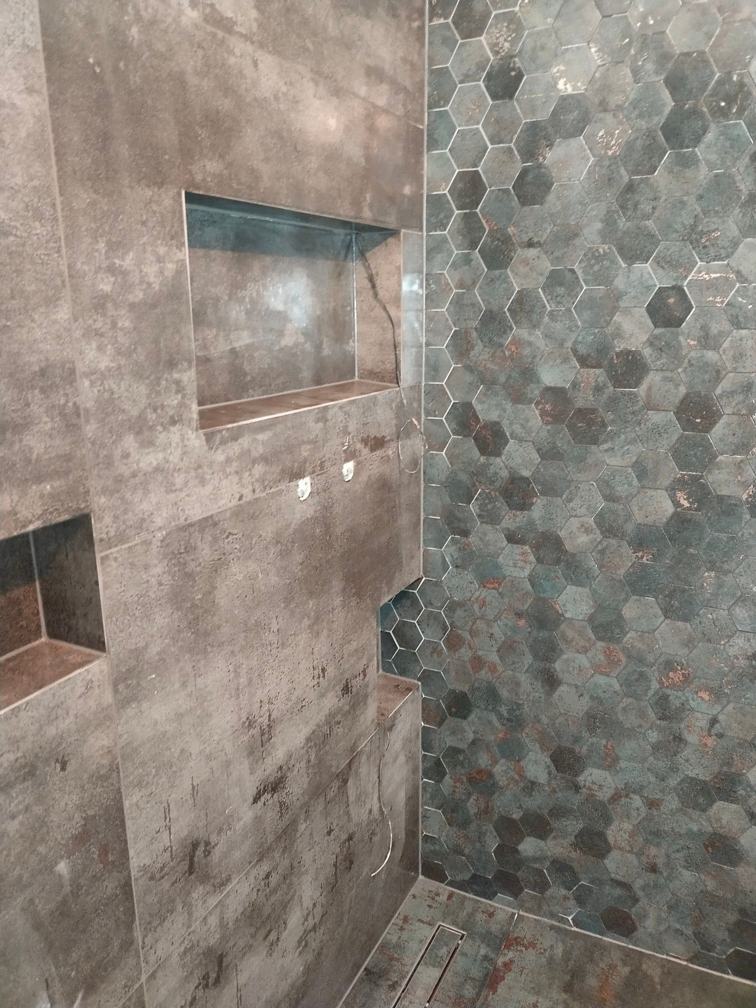 Wykończona ściana prysznicowa z wnękami i heksagonalnymi płytkami w odcieniach szarości i zieleni, widoczny odpływ liniowy.