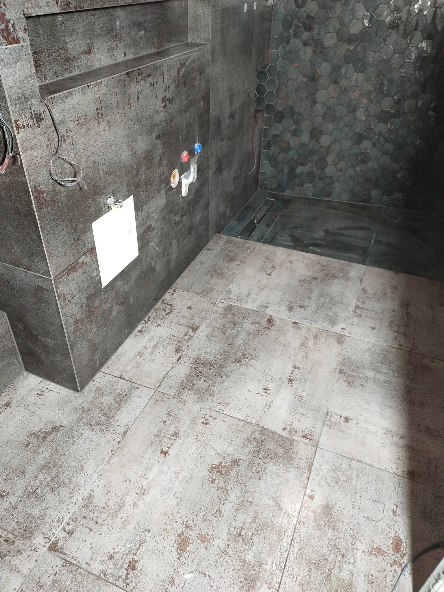 Wnętrze łazienki w trakcie prac wykończeniowych, widoczne szare płytki imitujące rdzę na ścianach i podłodze, heksagonalne płytki w strefie prysznica oraz wystające kable i instalacja wodna.