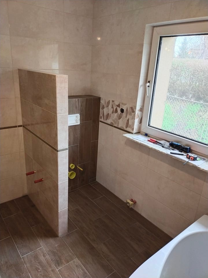 Wykończona łazienka z beżowymi płytkami ściennymi, brązowymi płytkami podłogowymi imitującymi drewno, zabudową WC i oknem z widokiem na zewnątrz. Na parapecie leżą narzędzia.