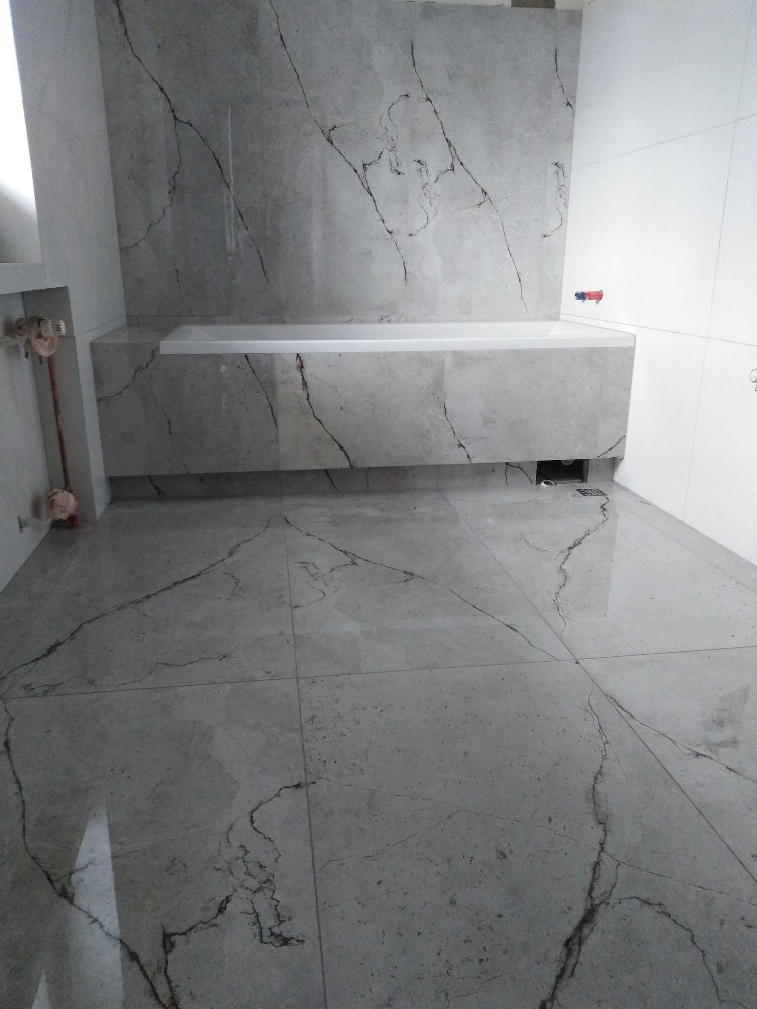 Wykończona łazienka z wanną obudowaną płytkami imitującymi marmur, podłoga wyłożona podobnymi płytkami, widoczny odpływ liniowy przy wannie.