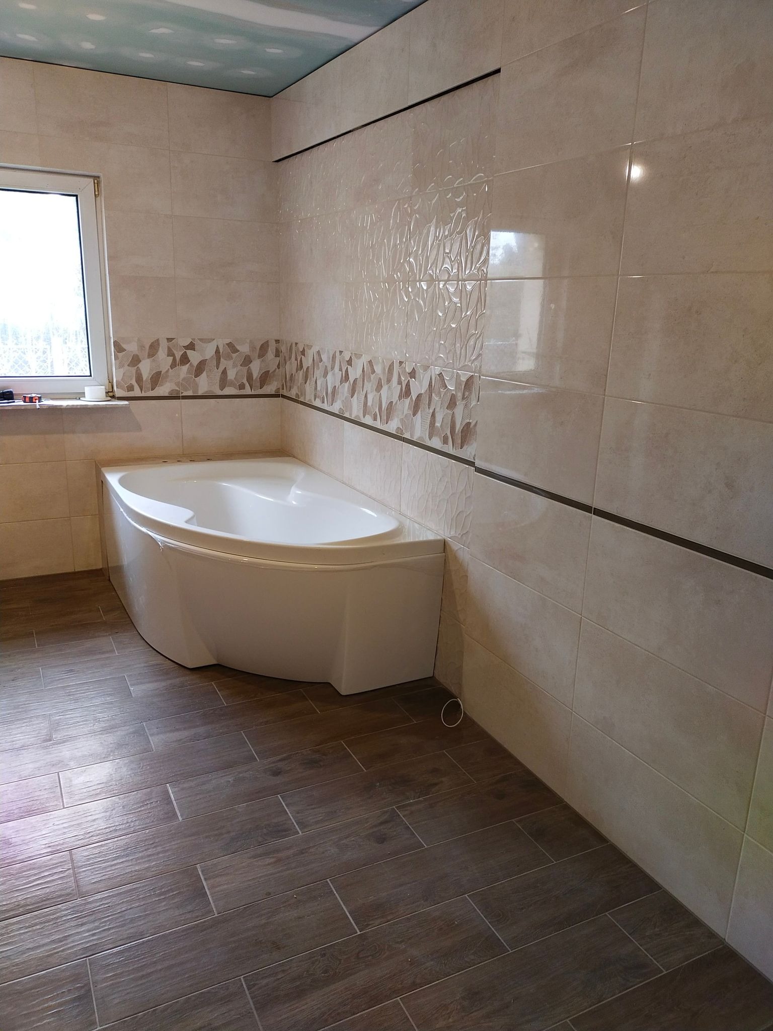 Jasna łazienka po remoncie z narożną wanną, brązowymi panelami podłogowymi imitującymi drewno i beżowymi płytkami ściennymi z dekoracyjnym pasem z motywem liści.