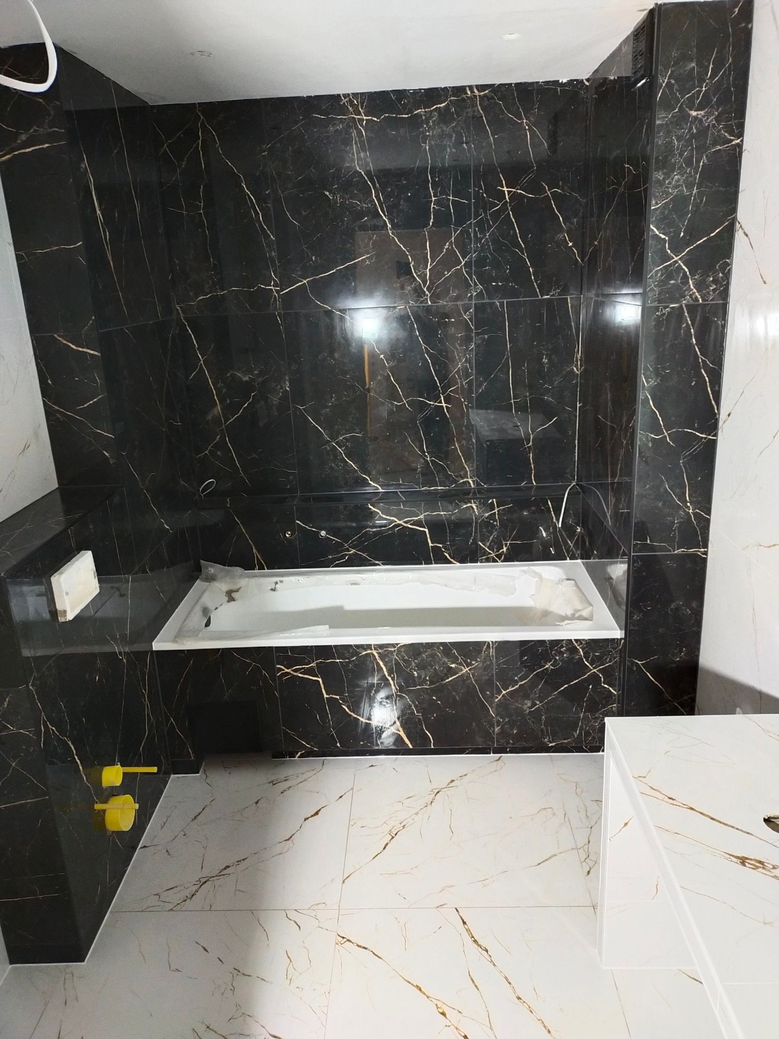 Nowoczesna łazienka z czarnymi płytkami imitującymi marmur ze złotymi żyłami na ścianach i białymi płytkami na podłodze, wanna w zabudowie, widoczne żółte narzędzia glazurnicze.