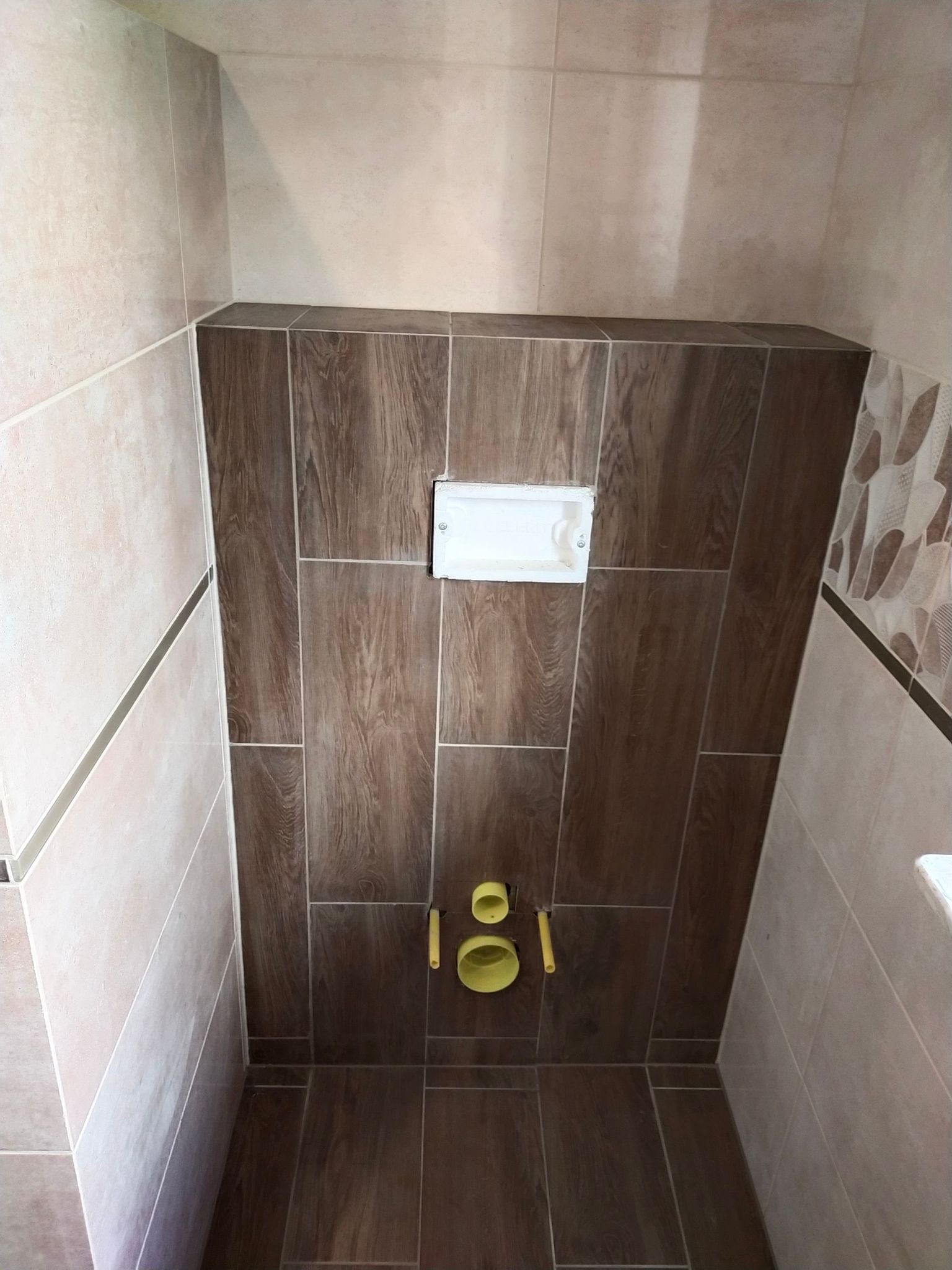 Świeżo wykończona toaleta w łazience, ściany i podłoga wyłożone płytkami imitującymi drewno, widoczny stelaż podtynkowy WC z przyciskiem spłukującym i rurami odpływowymi.