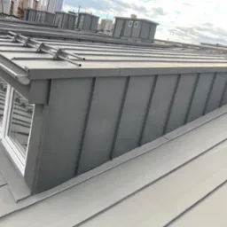 Szary dach z blachy trapezowej z oknem dachowym i rynnami, widok z góry na tle błękitnego nieba.