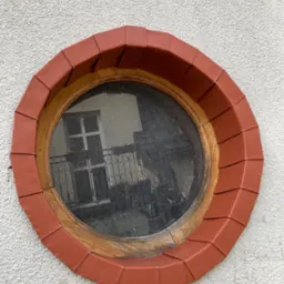Okrągłe okno w elewacji budynku, obramowane drewnem i ceramiczną, czerwoną dachówką, odbijające fragment budynku z balkonem.