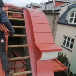 Dekarz w trakcie wymiany czerwonej dachówki na dachu budynku w zabudowie miejskiej, widoczna więźba dachowa i kominy w tle.