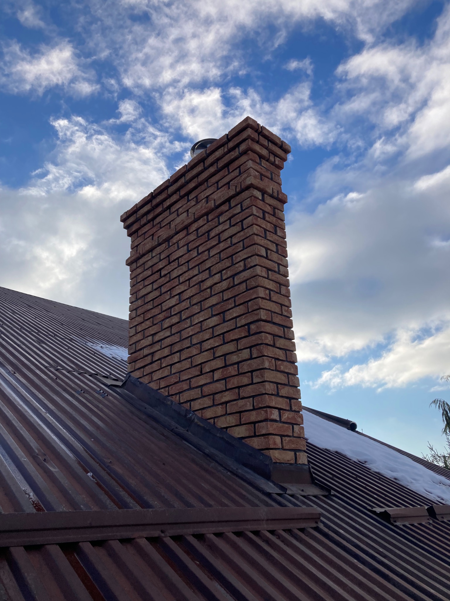 Ceglany komin na metalowym dachu z niewielką ilością śniegu, na tle błękitnego nieba z białymi obłokami.