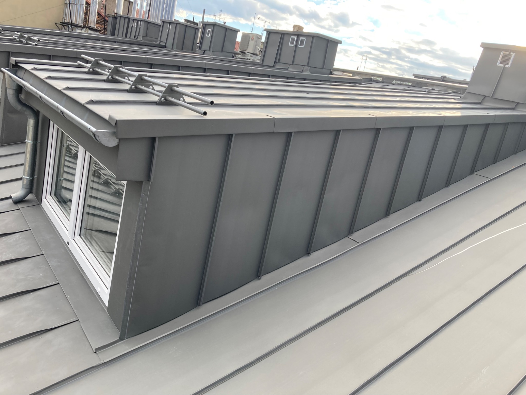 Szary dach z blachy trapezowej z oknem dachowym i rynnami, widok z góry na tle błękitnego nieba.