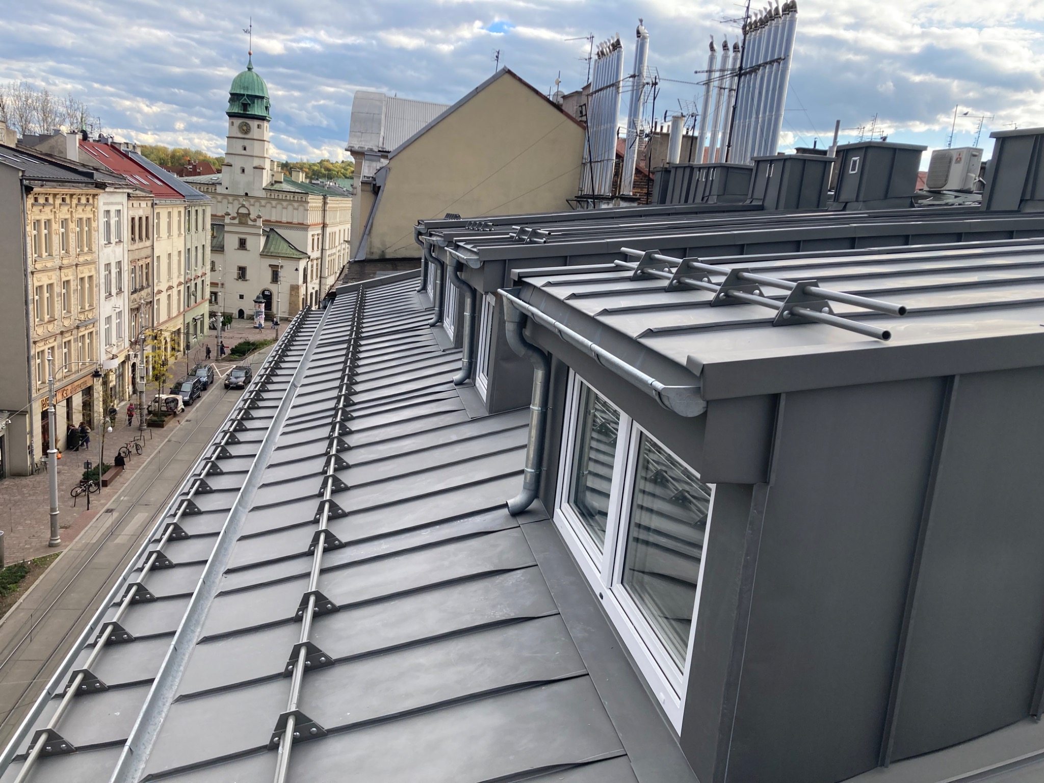 Nowoczesny, szary dach z blachy trapezowej z oknami dachowymi i systemem rynnowym, widok z góry na miejską zabudowę z wieżą ratuszową w tle.