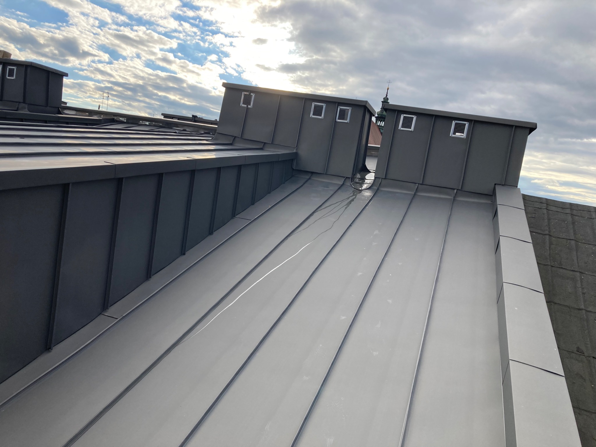 Szary, metalowy dach z kominami wentylacyjnymi na tle błękitnego nieba z chmurami, widok na wieżę kościoła w oddali.