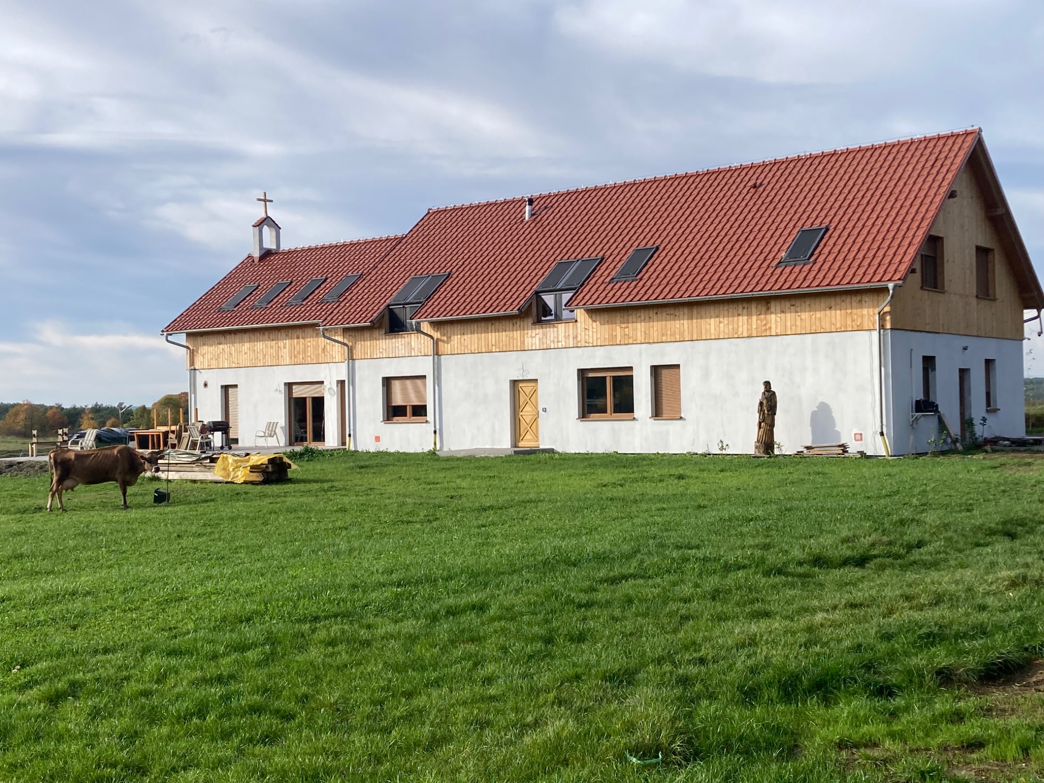 Nowo wybudowany dom z czerwoną dachówką i oknami dachowymi, z drewnianą elewacją na poddaszu, widziany z zewnątrz na tle zielonego trawnika i błękitnego nieba z krową na pierwszym planie.