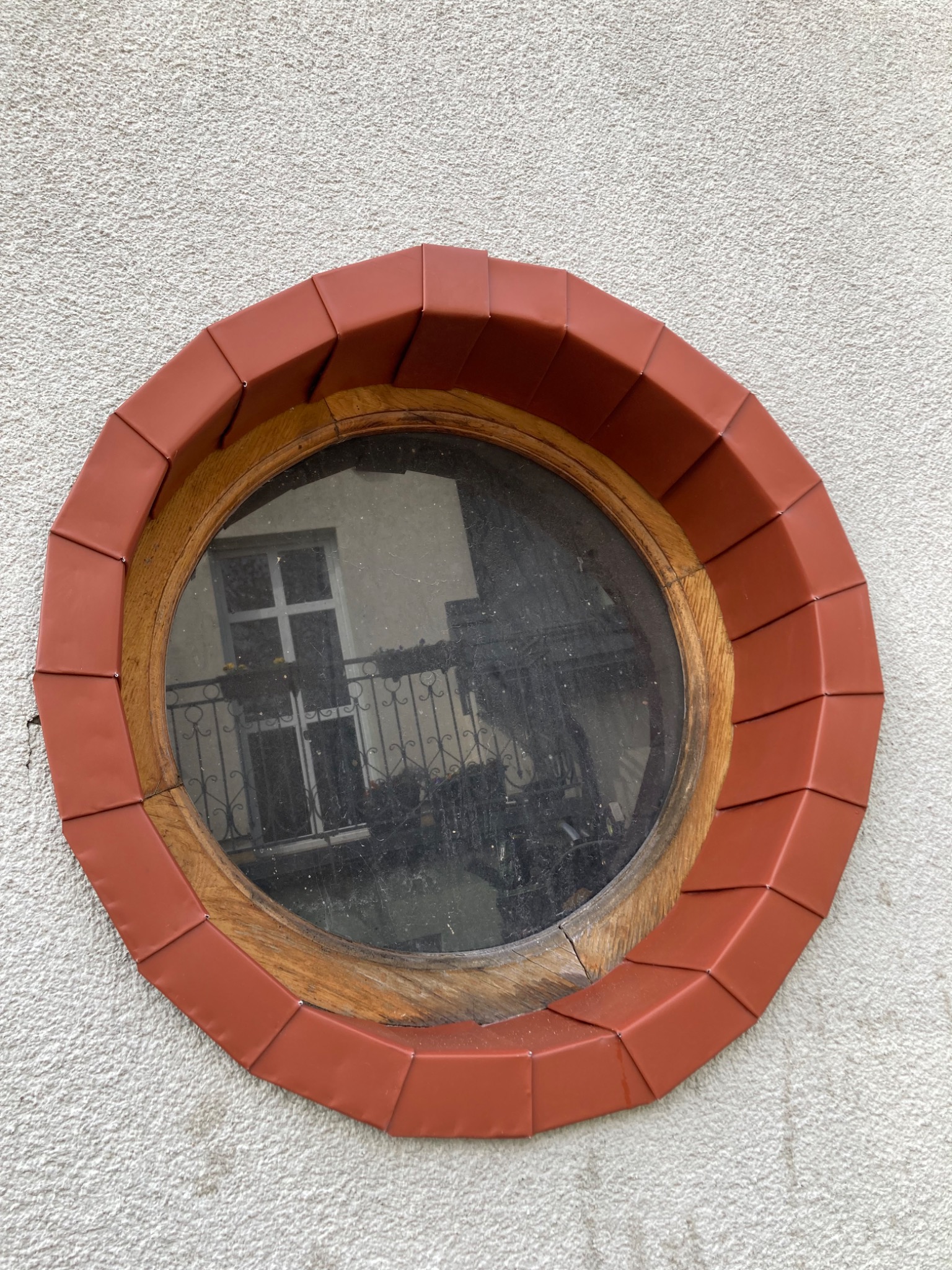 Okrągłe okno w elewacji budynku, obramowane drewnem i ceramiczną, czerwoną dachówką, odbijające fragment budynku z balkonem.