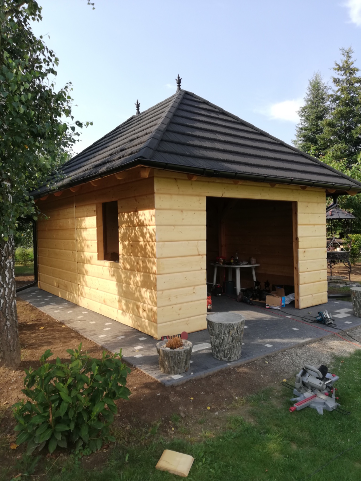 Drewniana altana z ciemnym dachem z dachówki, widoczne narzędzia i materiały budowlane, częściowo wykończony teren wokół.