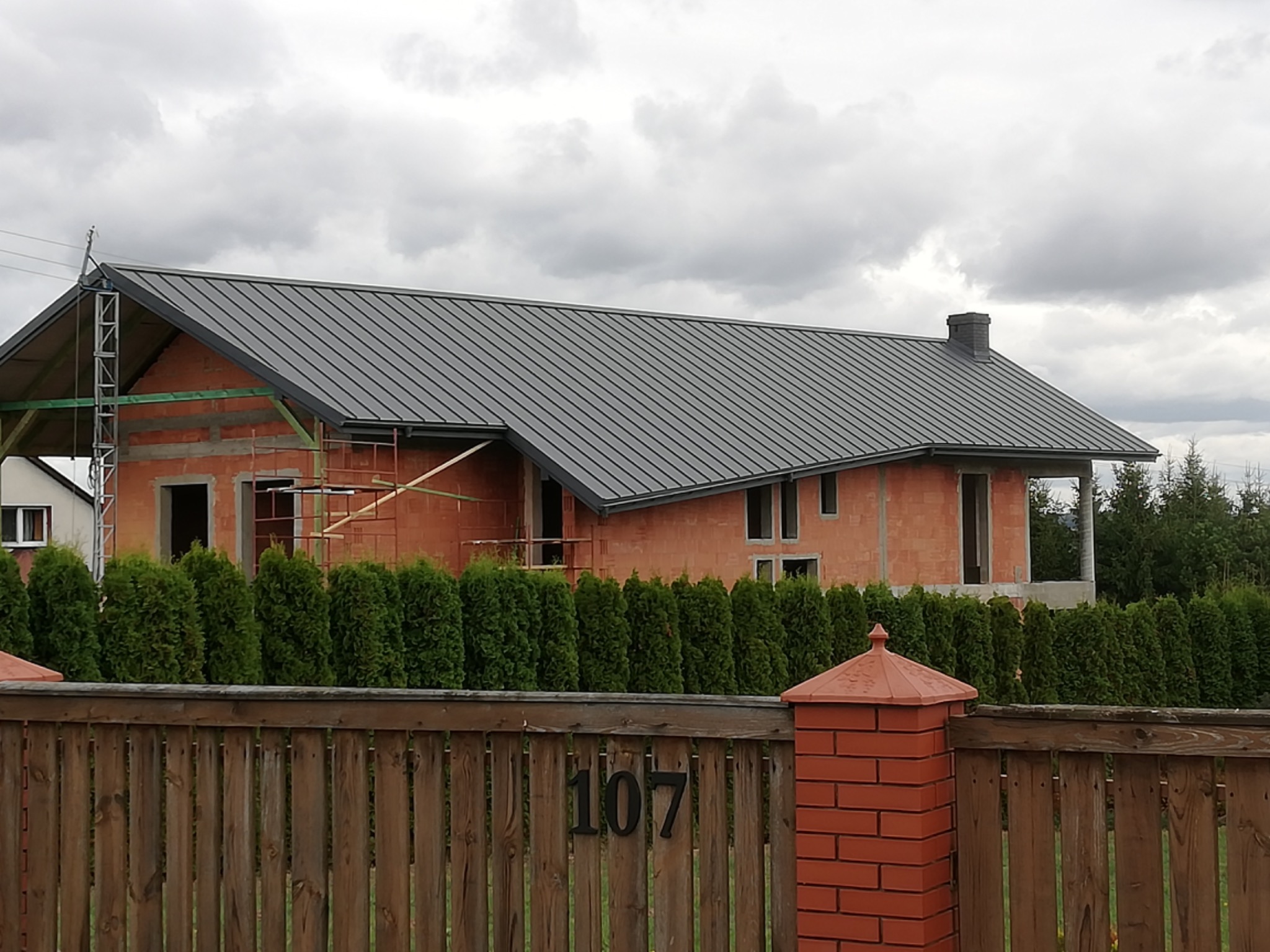 Dom w stanie surowym otwartym z nowym, szarym pokryciem w Miechowie, widoczny zza drewnianego płotu z numerem 107.